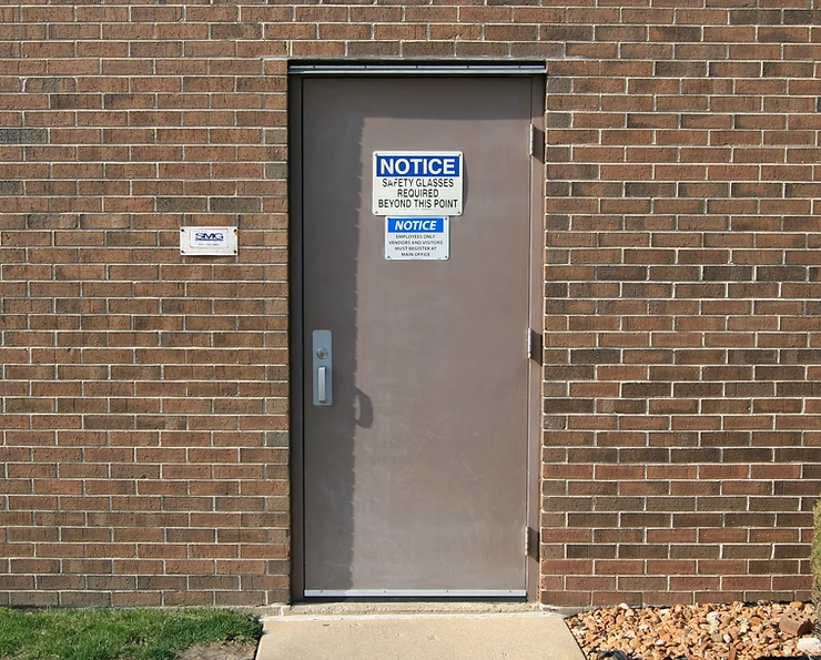 A brown, fire-rated metal door set in a brick wall displays multiple safety notice signs, including a prominent one instructing that safety glasses are required beyond the door. The area around the door includes a patch of green grass, some mulch with rocks, and a concrete step.