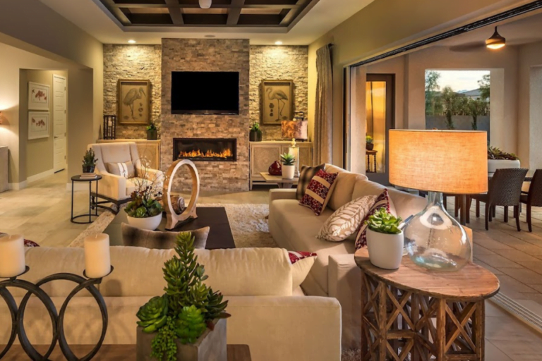 A cozy modern living room features a stone fireplace with a mounted TV above, surrounded by warm lighting. Cream-colored sofas are adorned with red and beige pillows. A glass door opens to a patio showcasing stylish outdoor fireplaces. Decorative plants and contemporary art pieces enhance the space.