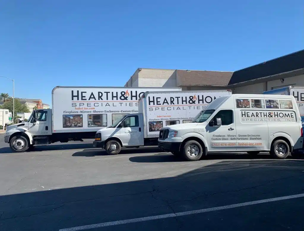 Three service vehicles marked with signs for "Hearth & Home Specialties." are parked in a lot. The vehicles include a large truck, a medium-sized van, and a smaller van. Each vehicle displays images and text promoting fireplaces, mirrors, shower enclosures, and custom glass—truly embodying our About Us vision.