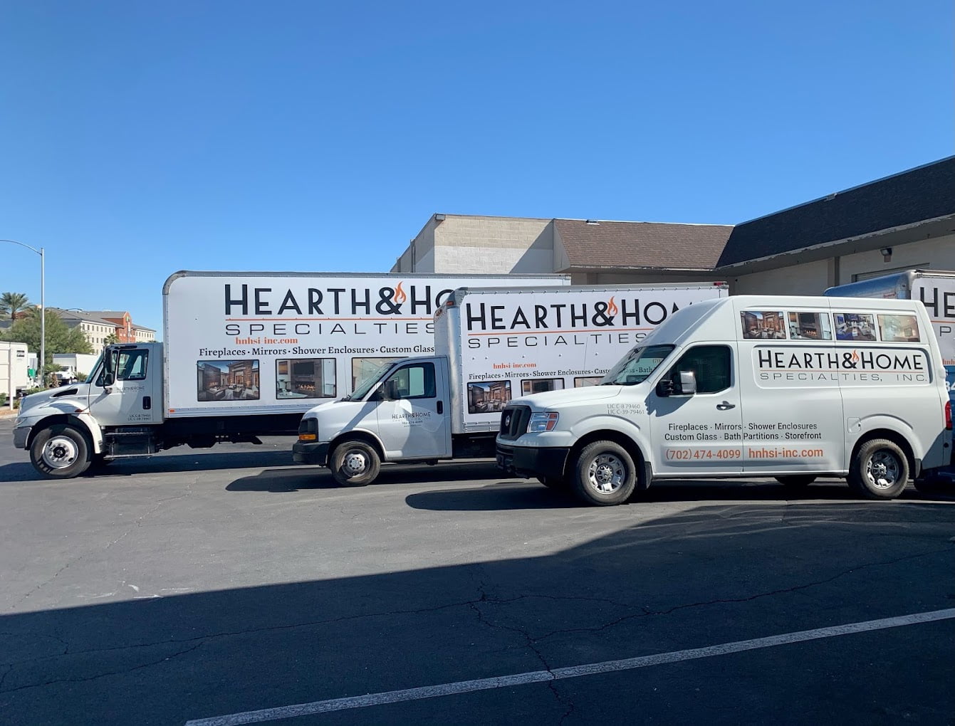 Three service vehicles marked with signs for "Hearth & Home Specialties, Inc." are parked in a lot. The vehicles include a large truck, a medium-sized van, and a smaller van. Each vehicle displays images and text promoting fireplaces, mirrors, shower enclosures, and custom glass—truly embodying our About Us vision.