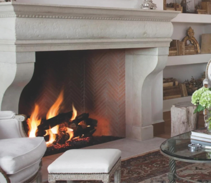 A cozy living room with a lit fireplace creates a warm atmosphere. The space features a light-colored mantel, upholstered armchairs, an ottoman, and a glass-top coffee table adorned with green hydrangeas. Built-in shelves filled with books and decorative items complete this outdoor-inspired haven.