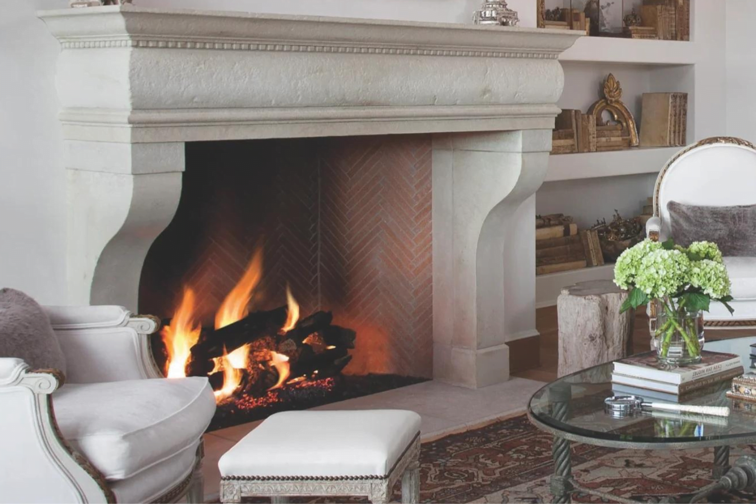 A cozy living room with a lit fireplace creates a warm atmosphere. The space features a light-colored mantel, upholstered armchairs, an ottoman, and a glass-top coffee table adorned with green hydrangeas. Built-in shelves filled with books and decorative items complete this outdoor-inspired haven.