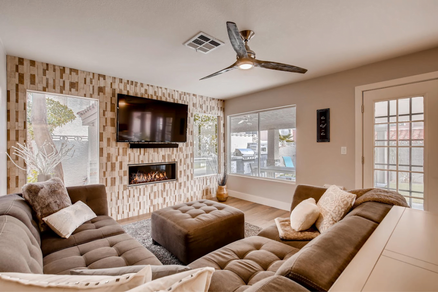 A modern living room features a sectional sofa, an ottoman, and a large flat-screen TV mounted above a fireplace with a patterned tile surround. Windows and a glass door provide natural light, revealing an inviting outdoor space. A ceiling fan hangs above.