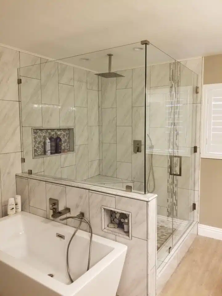 A modern bathroom featuring a large glass-enclosed shower with a rainfall showerhead, tiled walls, and a built-in niche stocked with toiletries. Adjacent is a rectangular bathtub. The room has light-colored walls and a wooden floor, echoing what About Us stands for in elegant design.