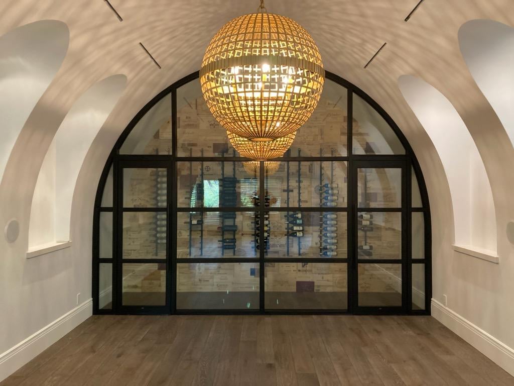 The image shows a room with a large, arched entrance framed by black metal and glass doors. Inside, there is a modern wine cellar with wooden shelves lined with wine bottles. A large, round, woven light fixture hangs from the ceiling, illuminating the space. Learn more about us and our exquisite designs.