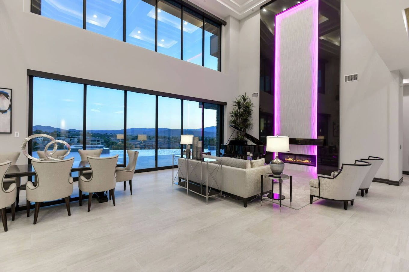 A modern, luxurious living area features a large glass wall offering expansive views of mountains and an outdoor pool. The room, crafted by Hearth & Home Specialties, is decorated with contemporary furniture, including a dining table, comfortable sofas, and two armchairs. A tall, illuminated pink accent wall houses a sleek fireplace.