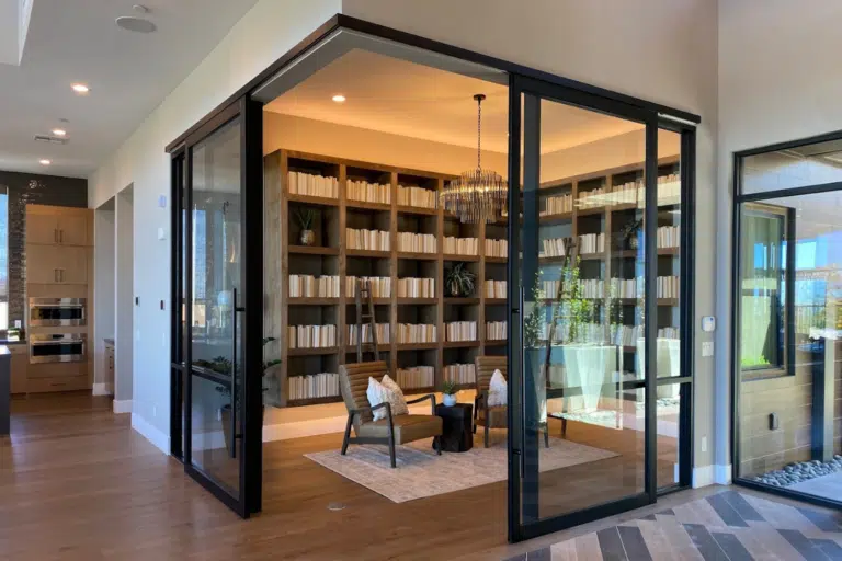 A modern room with custom glass walls features a cozy seating area with two chairs and a rug. Shelves filled with books line the back wall, and a chandelier hangs from the ceiling. Bright with natural light and warm wood flooring, this space embodies Hearth & Home Specialties.