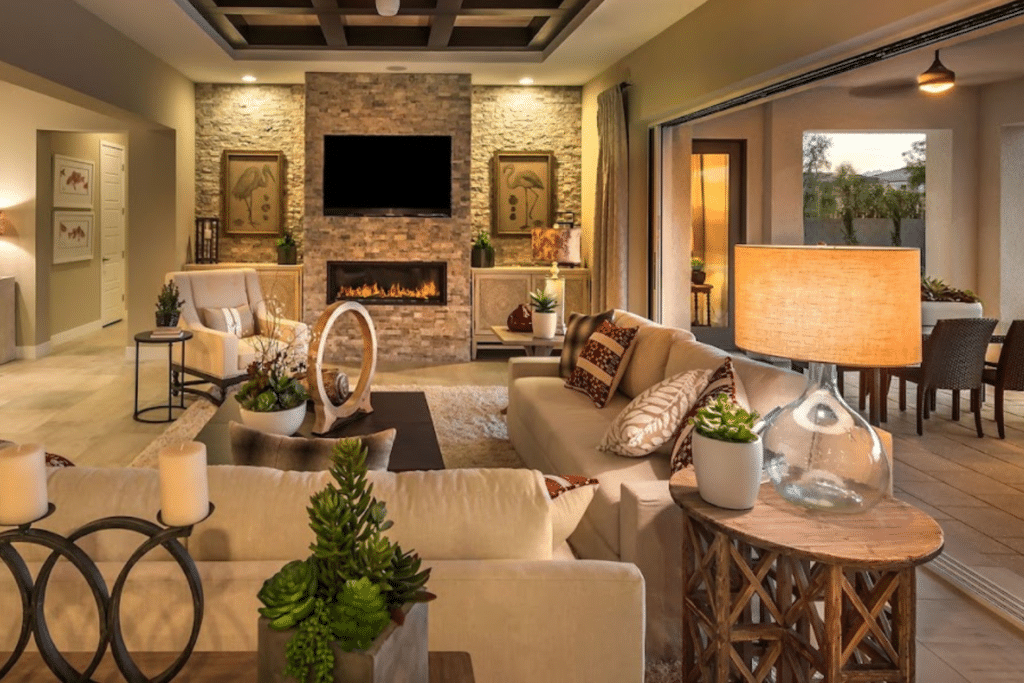 A cozy living room features a modern gas fireplace with a large TV above and stylish decor. Cream sofas and a wooden coffee table sit on a rug. Sliding custom glass doors open to an outdoor dining area, flooding the space with natural light. Earthy tones create a warm ambiance.