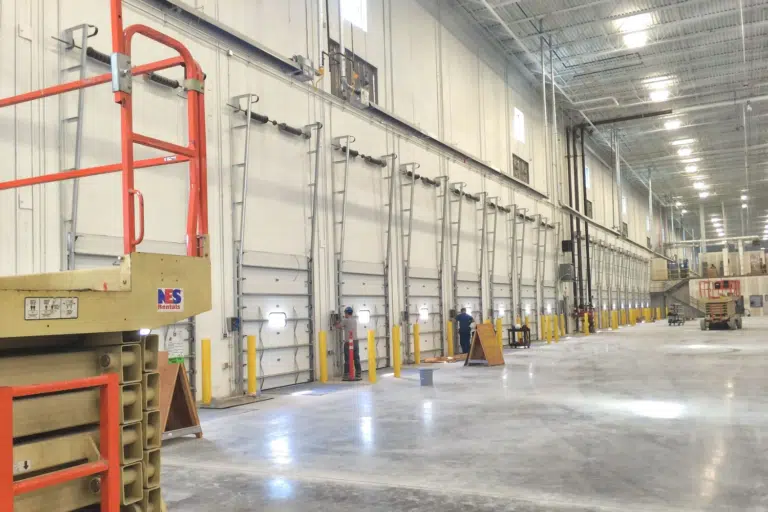 The interior of the large warehouse, belonging to Hearth & Home Specialties in Las Vegas, features multiple loading dock doors along one wall. A scissor lift is visible in the foreground on the left, while a few workers are seen near the doors in the distance. The space is well-lit with a high ceiling.