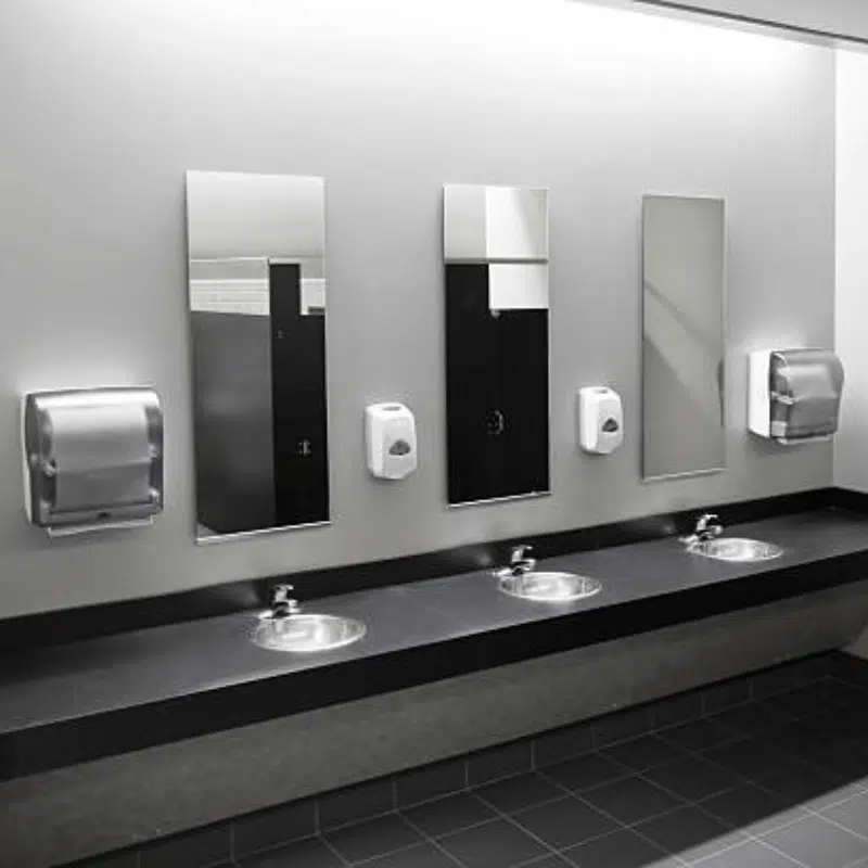 A modern public restroom, designed with commercial services in mind, features three sinks with individual mirrors and soap dispensers mounted on the wall. Paper towel dispensers are located on either end above the counters. The floor is tiled in a dark color, creating a minimalist yet functional space.