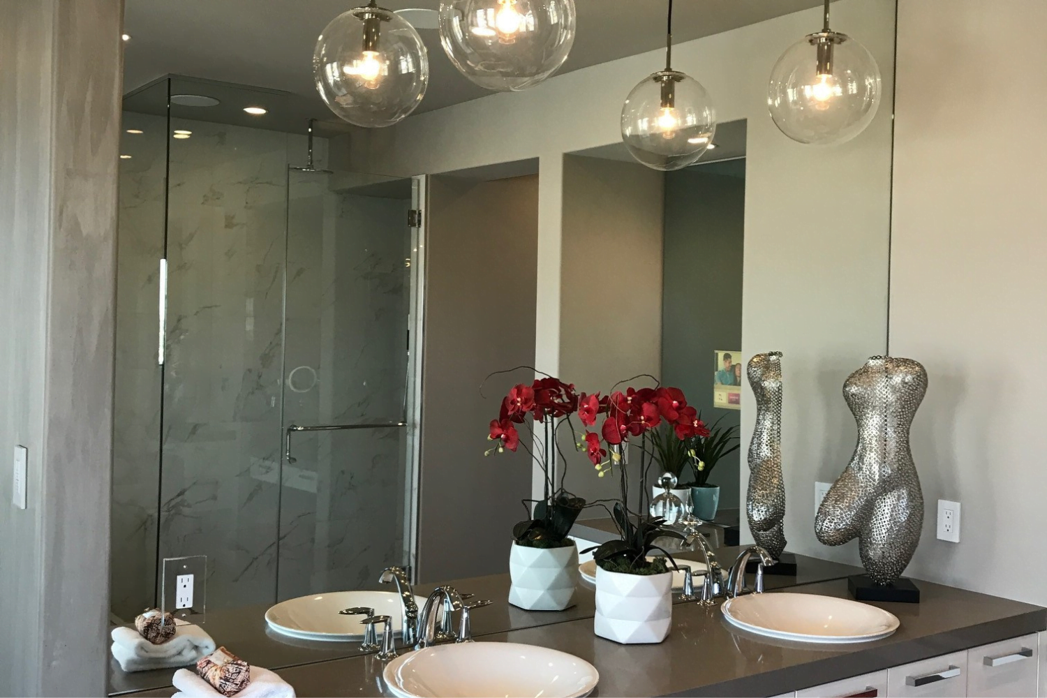 A modern bathroom with a large mirror, expertly installed by Custom Glass Services, faces two sinks with silver faucets. Each sink is accompanied by a white hexagonal pot containing red orchids. Above the sinks are three spherical pendant lights. To the right, there are decorative silver statues of torsos.