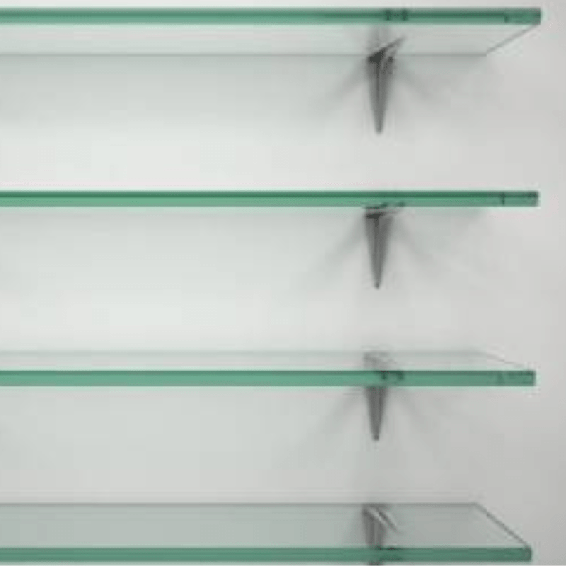 Four empty glass shelves, crafted through Custom Glass Services, are mounted on a white wall, supported by metallic brackets. The shelves are evenly spaced and exude a clean, minimalist aesthetic.