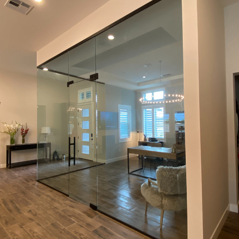 A modern room with custom glass walls and wooden flooring features a desk, chair, and fluffy beige rug. The room has a lot of natural light coming through large windows with blinds. A hallway adorned with decorative pieces can be seen through the glass wall on the left.