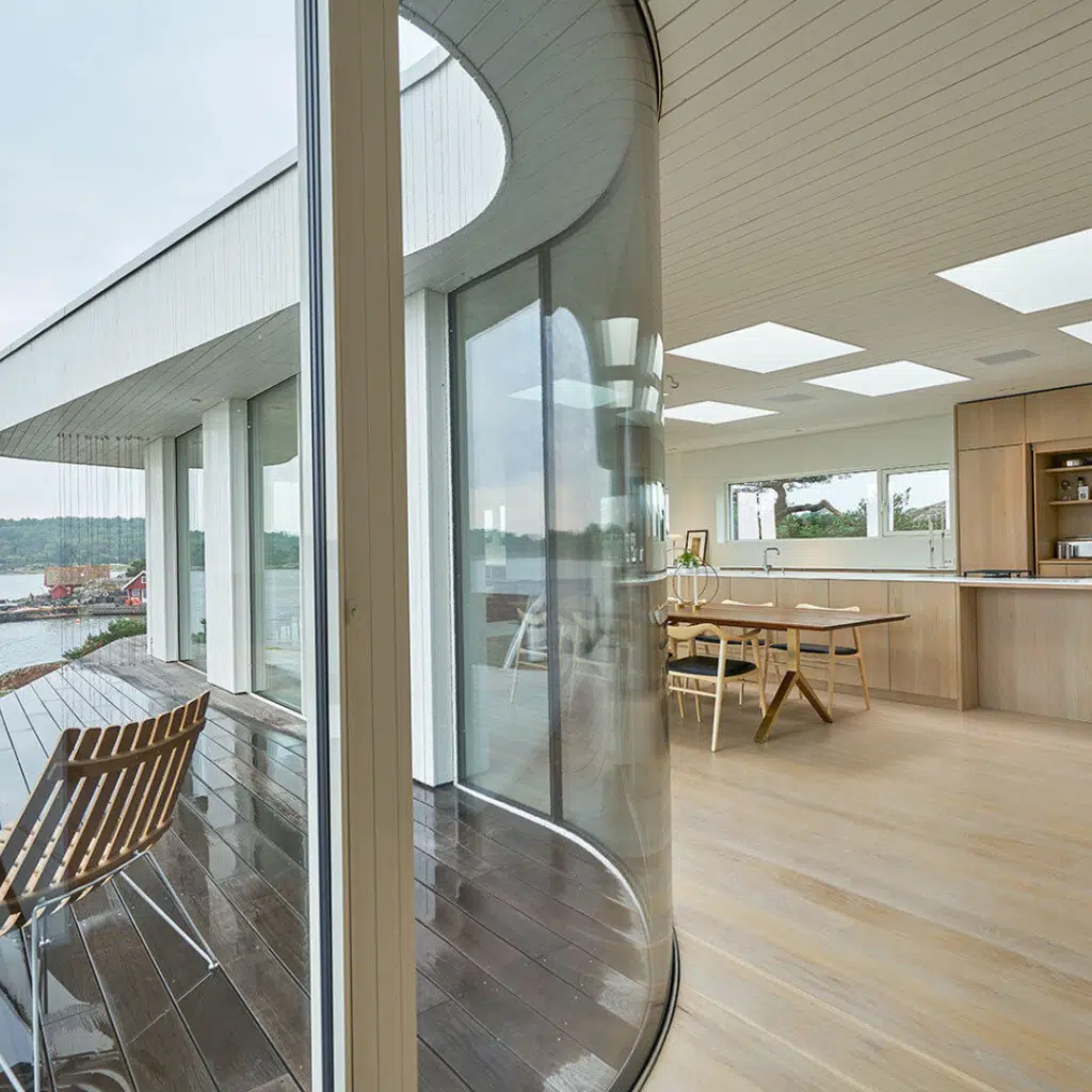 Modern interior with large curved custom glass walls, wooden flooring, and an open kitchen. A small balcony with seating overlooks a scenic outdoor view. Natural light floods the space through ceiling windows and expansive glass panes, offering a warm hearth & home ambiance.