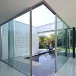 A modern interior courtyard in Las Vegas boasts large glass walls and a rectangular reflecting pool. A person sits by the pool, enveloped in minimalistic, white architecture with green garden views, showcasing Hearth & Home Specialties' elegant design approach for commercial spaces.