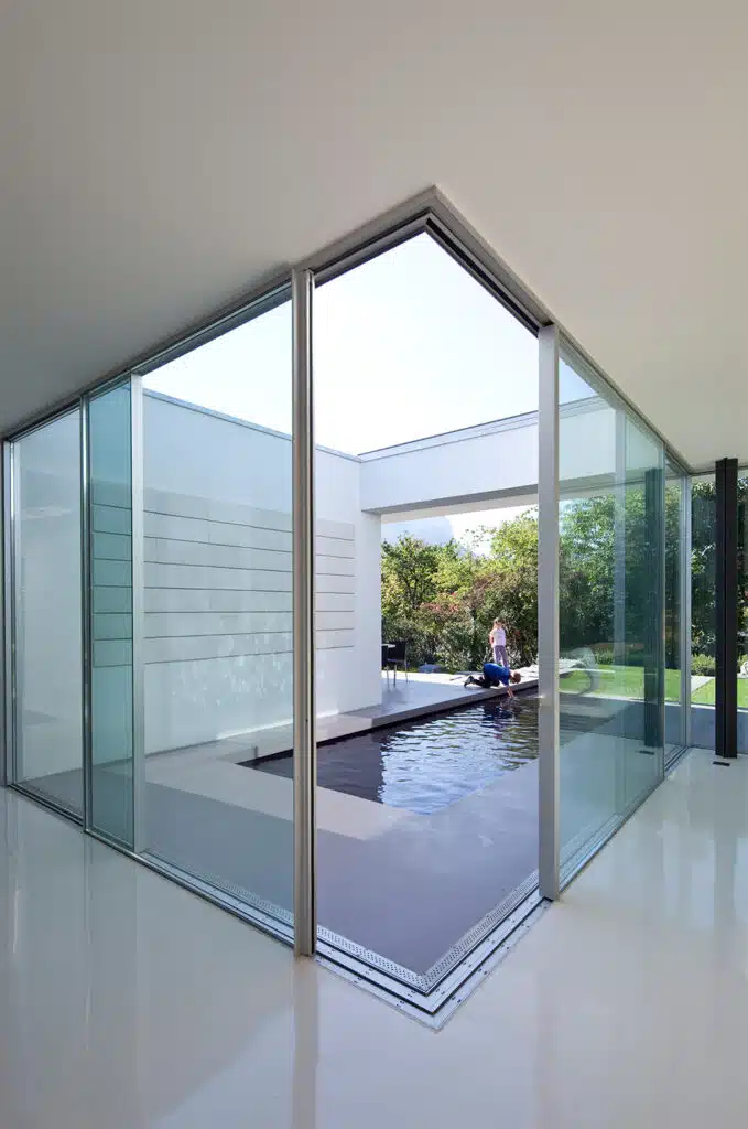A modern interior courtyard in Las Vegas boasts large glass walls and a rectangular reflecting pool. A person sits by the pool, enveloped in minimalistic, white architecture with green garden views, showcasing Hearth & Home Specialties' elegant design approach for commercial spaces.