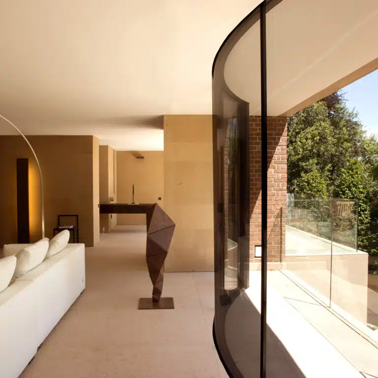 A modern living room with beige walls, a white sofa, and a geometric sculpture. Large curved glass windows offer a view of lush greenery outside. The room exudes warmth, accented by a sleek gas fireplace that complements its minimalist decor and earthy tones.