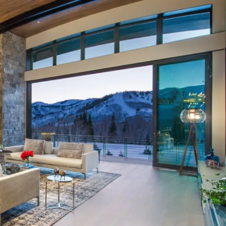 A modern living room with high ceilings and large windows offers a stunning view of snow-covered mountains. The room, designed by Hearth & Home Specialties, features beige sofas, a glass coffee table with a red flower centerpiece, and a tall lamp beside the elegant gas fireplace for a cozy, contemporary feel.