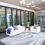 Modern showroom with large glass panel doors and windows on display. Two white sofas with blue and grey pillows are placed on a patterned rug. A small glass coffee table with a plant and brochure sits between the sofas. Overhead lighting features round bulbs, complementing the elegant gas fireplaces showcased nearby.