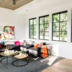 Bright living room with large windows and a modern black sectional sofa adorned with colorful cushions. An abstract painting graces the wall above a sleek custom glass fireplace. A set of round coffee tables with candles accents the space, while light wood flooring and a striped wood ceiling complete the look.