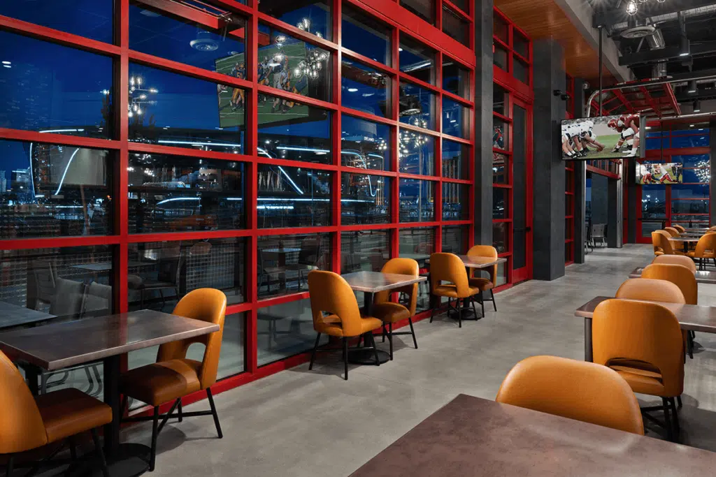 A modern restaurant with red-framed floor-to-ceiling windows features cozy gas fireplaces. Orange chairs and dark tables are neatly arranged. Several TVs display sports channels. Outside, the city skyline is visible under a night sky. The ambiance is sleek and inviting.
