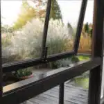 A partially open black-framed window reveals a view of a lush garden with tall grasses and trees under soft sunlight, accompanied by the cozy warmth of nearby gas fireplaces. A wooden deck is visible outside.
