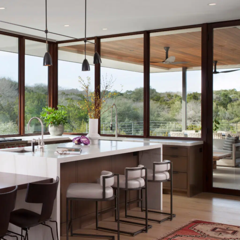 Modern kitchen with large windows offering a view of lush greenery. Features a white marble island with barstools, pendant lighting, and wooden cabinetry. A custom glass partition elegantly separates the space. A red rug adds warmth, while a vase with branches and green plants decorates the counter.
