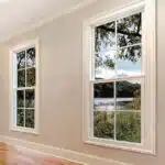 A room with two large windows, showcasing a scenic view of a river, trees, and a cloudy sky. The space features light-colored walls and wooden flooring, complemented by a custom glass design that enhances its elegance.