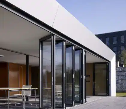 Modern building with custom glass doors opening to a wooden deck. The interior has minimalistic furnishings, and sunlight streams in, creating a bright and airy atmosphere. The structure features clean lines and sleek design elements, complemented by chic fireplaces perfect for Las Vegas living.