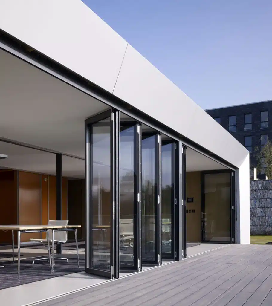 Modern building with custom glass doors opening to a wooden deck. The interior has minimalistic furnishings, and sunlight streams in, creating a bright and airy atmosphere. The structure features clean lines and sleek design elements, complemented by chic fireplaces perfect for Las Vegas living.