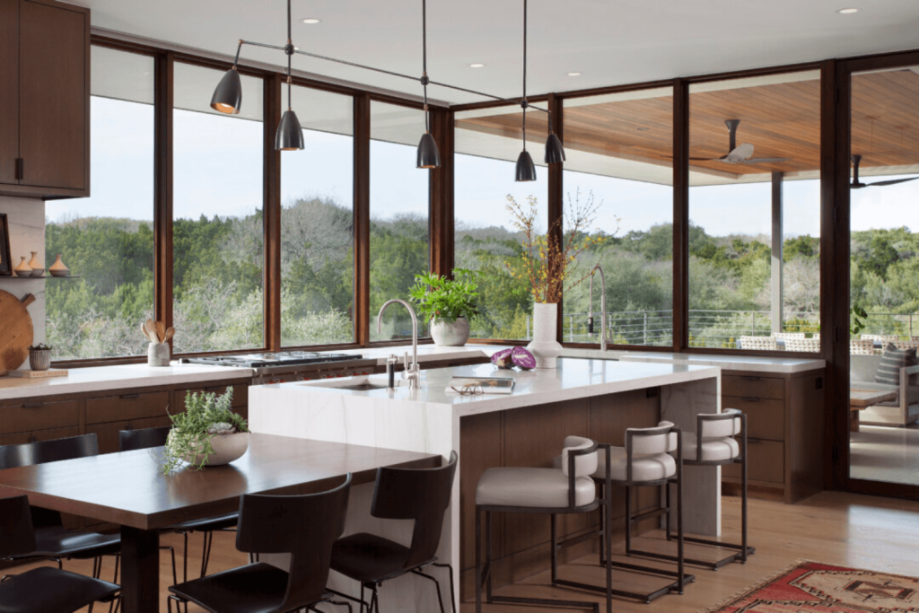 Spacious kitchen with large windows offering a scenic view of trees. It features a white marble island with bar stools by Hearth & Home Specialties, black pendant lights, a dining table with black chairs, and wooden cabinets. Ample natural light brightens the space.
