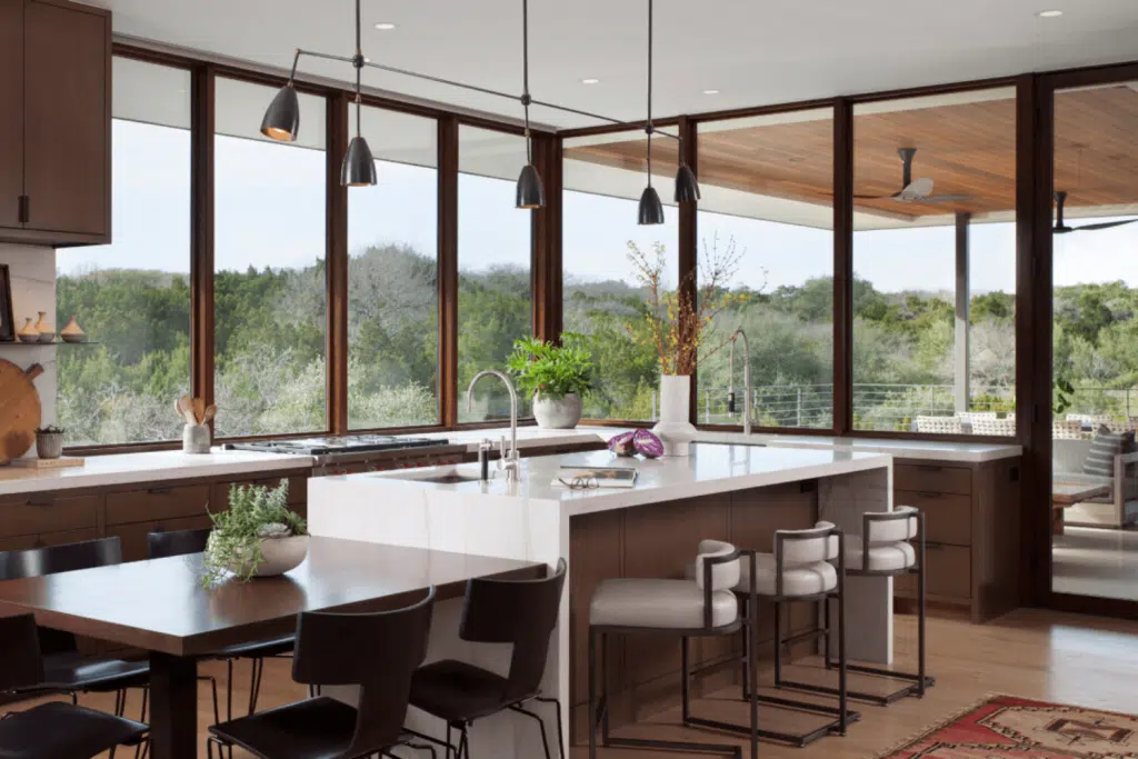 Spacious kitchen with large windows offering a scenic view of trees. It features a white marble island with bar stools by Hearth & Home Specialties, black pendant lights, a dining table with black chairs, and wooden cabinets. Ample natural light brightens the space.