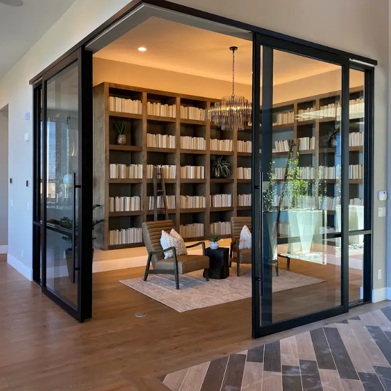 A modern home library with large glass sliding doors crafted by Custom Glass Services, a wooden floor, and built-in bookshelves filled with numerous books. Two cushioned armchairs with a small table between them sit on a rug in the center. A chandelier hangs from the ceiling.