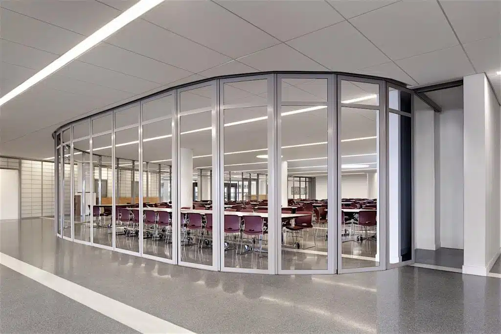 A modern, commercial classroom with floor-to-ceiling glass walls and a curved design. Inside are rows of tables and maroon chairs. The room is well-lit with overhead lighting and has a polished concrete floor, showcasing the sleek aesthetics of Hearth & Home Specialties.