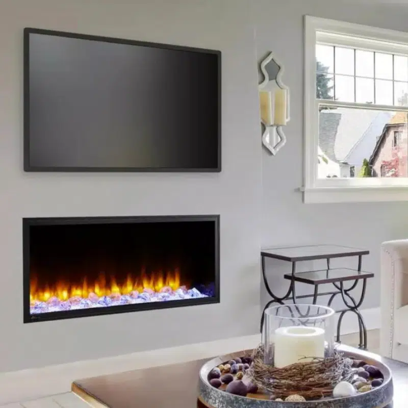 A modern living room showcases a wall-mounted TV above sleek fireplaces. A window with a view of rooftops graces the right, alongside a stylish mirror and a decorative side table adorned with a large candle and a bowl of ornaments.