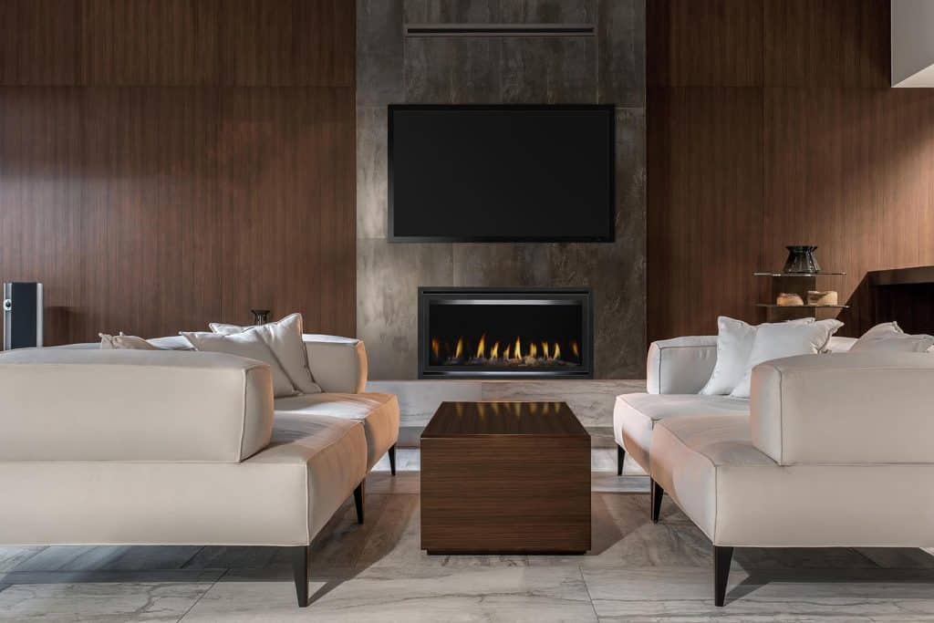 A modern living room with two beige sofas facing each other on a marble floor. A sleek wooden coffee table sits between them. A flat-screen TV and a fireplace are mounted on a dark wood-paneled wall. Minimalist decor creates a cozy atmosphere.