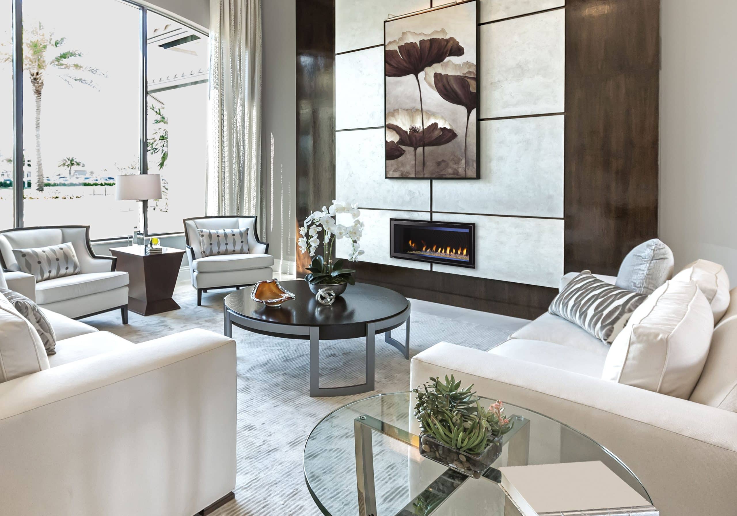 A modern living room with white sofas and armchairs around a round coffee table. A fireplace is set into a large wall, above which hangs a floral painting. Large windows provide natural light, and elegant decor includes plants and a lamp on a side table.