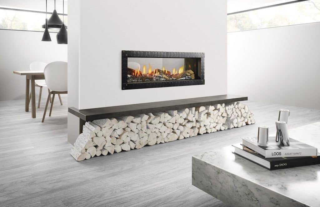 A modern living room with a sleek, rectangular fireplace embedded in a white wall. Below, neatly stacked firewood contrasts with the minimalist decor. A marble coffee table holds books and cups, and a dining area is visible in the background.