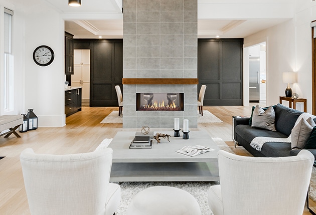 Modern living room with a central dual-sided fireplace. Neutral-toned furniture includes white chairs and a gray sofa with dark cushions. There are decorative elements on the coffee table and a cozy dining area visible in the background.