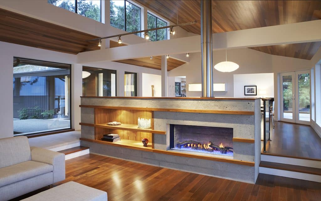 Modern living room with a central fireplace, surrounded by a concrete wall with shelves. Wooden ceiling and floors add warmth. Large windows offer natural light and a view of the outdoors. Minimalist decor with neutral tones and contemporary furniture.