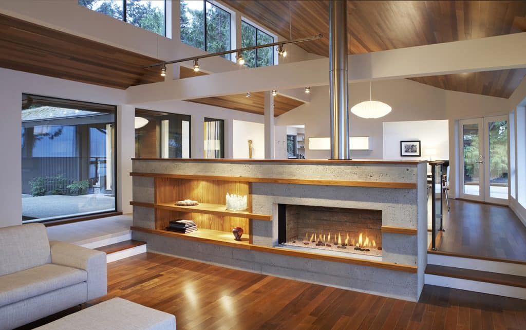 A modern living room featuring a central fireplace with open shelving, surrounded by wooden floors and ceiling. Large windows provide natural light, with a beige sofa and minimal decor completing the spacious, contemporary design.