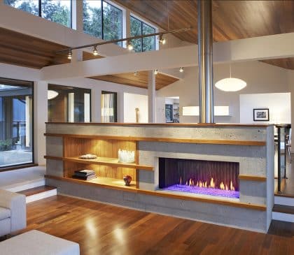Modern living room with wooden floors and ceiling, featuring a central concrete fireplace with blue flames. Shelves are built into the fireplace structure, and large windows provide natural light, offering a view of the outdoors.