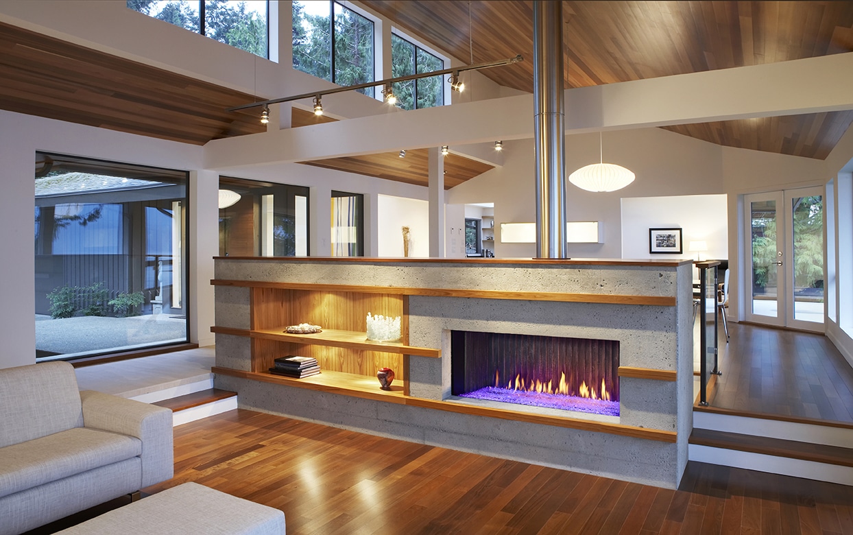 Modern living room with wooden floors and ceiling, featuring a central concrete fireplace with blue flames. Shelves are built into the fireplace structure, and large windows provide natural light, offering a view of the outdoors.