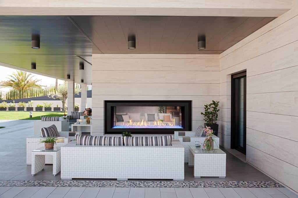 Outdoor patio featuring modern wicker furniture with striped cushions, a sleek gas fireplace set in a light stone wall, and potted plants. The covered area opens to a spacious yard with trees and a neatly landscaped garden.