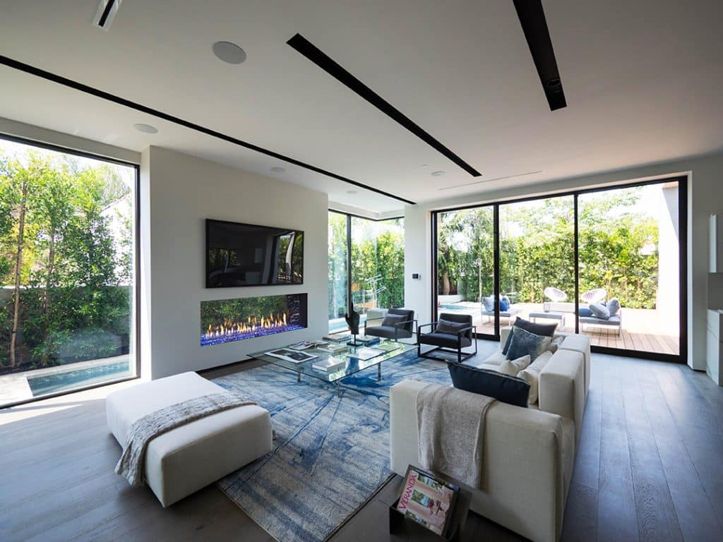 Spacious modern living room with large glass windows overlooking a garden. Features include a wall-mounted TV above a sleek fireplace, white sofas, a blue abstract rug, and a glass coffee table. Sliding doors open to an outdoor patio with seating.