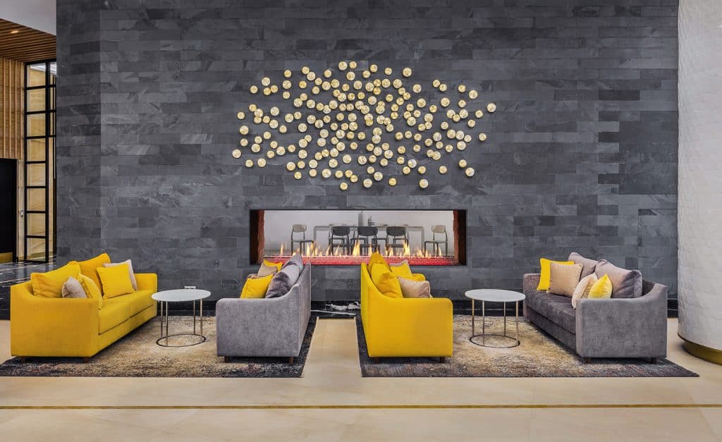 Modern lounge area with two grey sofas and two yellow sofas facing each other, centered around small round tables. A large wall with a decorative art installation and a linear fireplace enhances the contemporary ambiance.