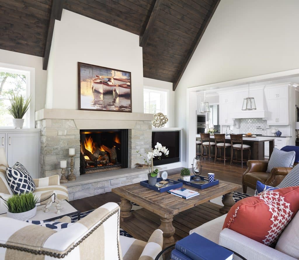 A cozy living room with a high ceiling features a stone fireplace, a wooden coffee table, and colorful patterned chairs. A painting of boats hangs above the mantel. In the background, a kitchen with a dining area is visible.