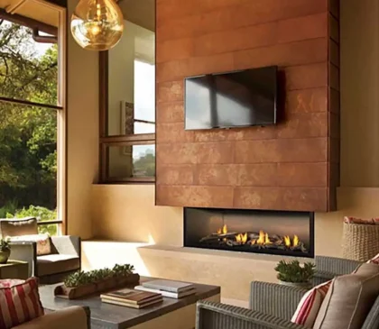 Cozy living room with large windows offering a garden view. A modern indoor fireplace sits beneath a mounted TV on a textured wall. The space features a sofa with striped cushions, a coffee table filled with books, potted plants, and a hanging glass light fixture.