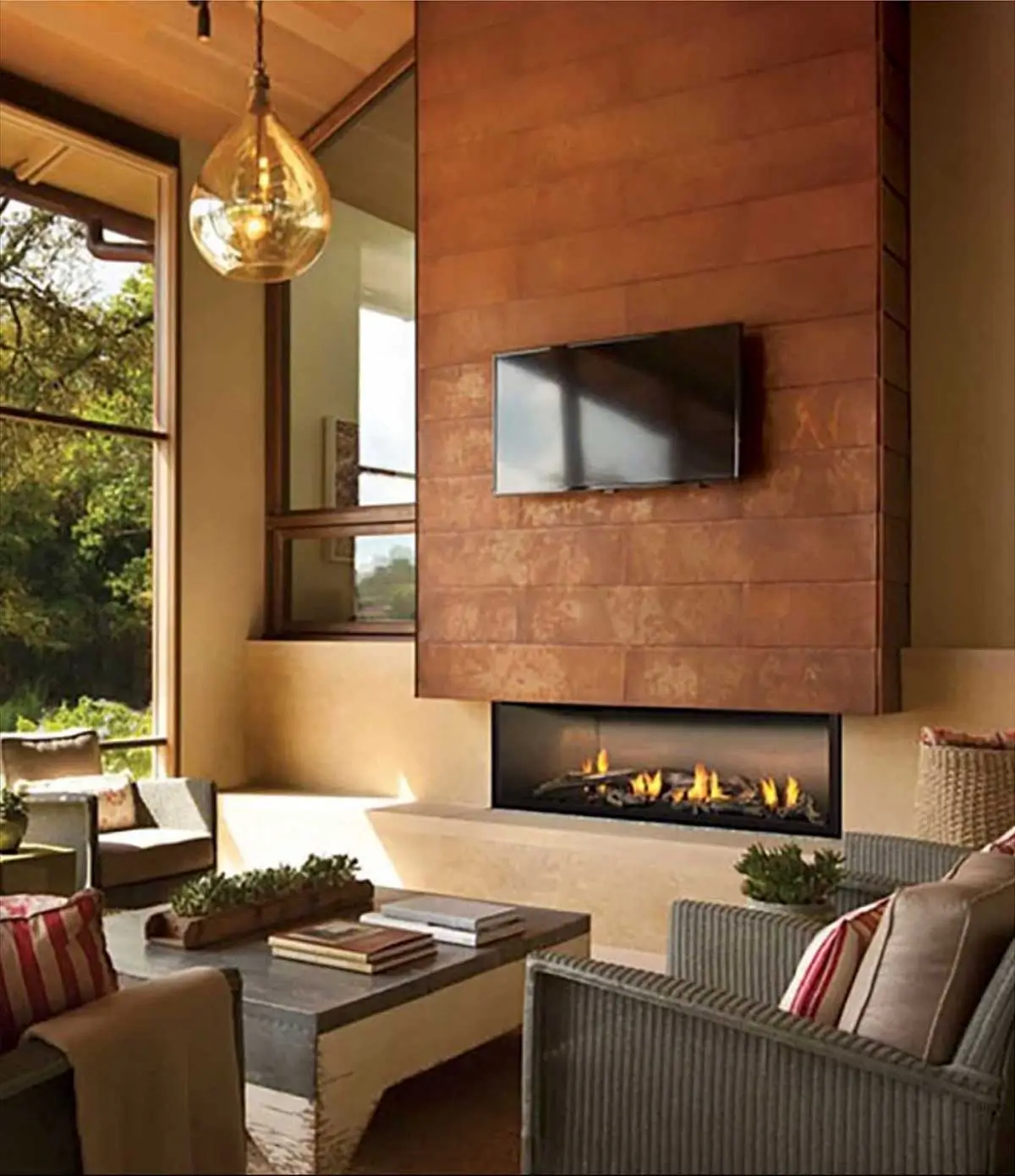 Cozy living room with large windows offering a garden view. A modern indoor fireplace sits beneath a mounted TV on a textured wall. The space features a sofa with striped cushions, a coffee table filled with books, potted plants, and a hanging glass light fixture.
