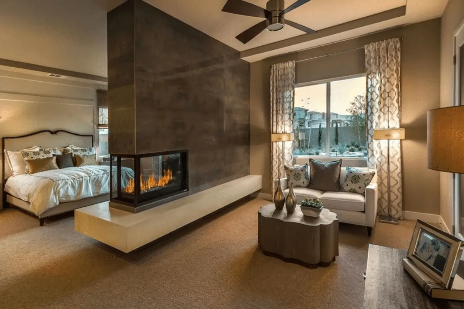 A modern bedroom and sitting area, separated by a double-sided fireplace from Hearth & Home Specialties. The room features neutral tones, a large window with curtains, a cushioned chair, a coffee table, and soft ambient lighting.